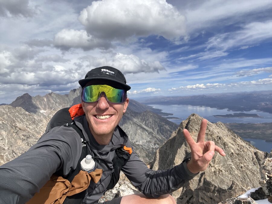 Nate Bowser on a summit in the Tetons, smiling, with mountains and a lake behind him.
